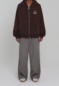 Sweat à capuche marron à fermeture éclair avec une grande capuche et un logo sur la poitrine, associé à un pantalon gris ample et des baskets blanches avec des semelles marron.