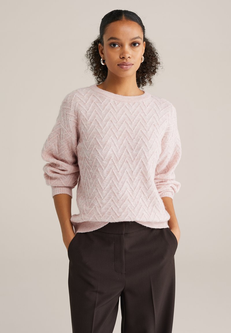 Világos rózsaszín kötött pulóver cikkcakk mintával, lekerekített nyakkivágással és felhajtott ujjal, sötétbarna nadrággal párosítva.