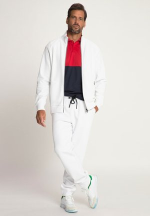 Chaqueta blanca con cremallera sobre una camiseta de polo en colores rojo y azul marino, combinada con pantalones de chándal blancos y zapatillas blancas con detalles verdes.