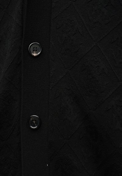 Μαύρο πλεκτό ύφασμα με διακριτικό μοτίβο διαμαντιού και λογότυπο σε τονικούς τόνους, με δύο μαύρα κουμπιά με τέσσερις οπές σε κάθετα μανίκια.