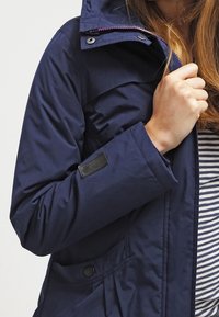 Giacca impermeabile di colore blu navy, con cappuccio, zip frontale e tasche con bottone. Presenta una fodera interna a righe e una toppa con logo sulla manica.