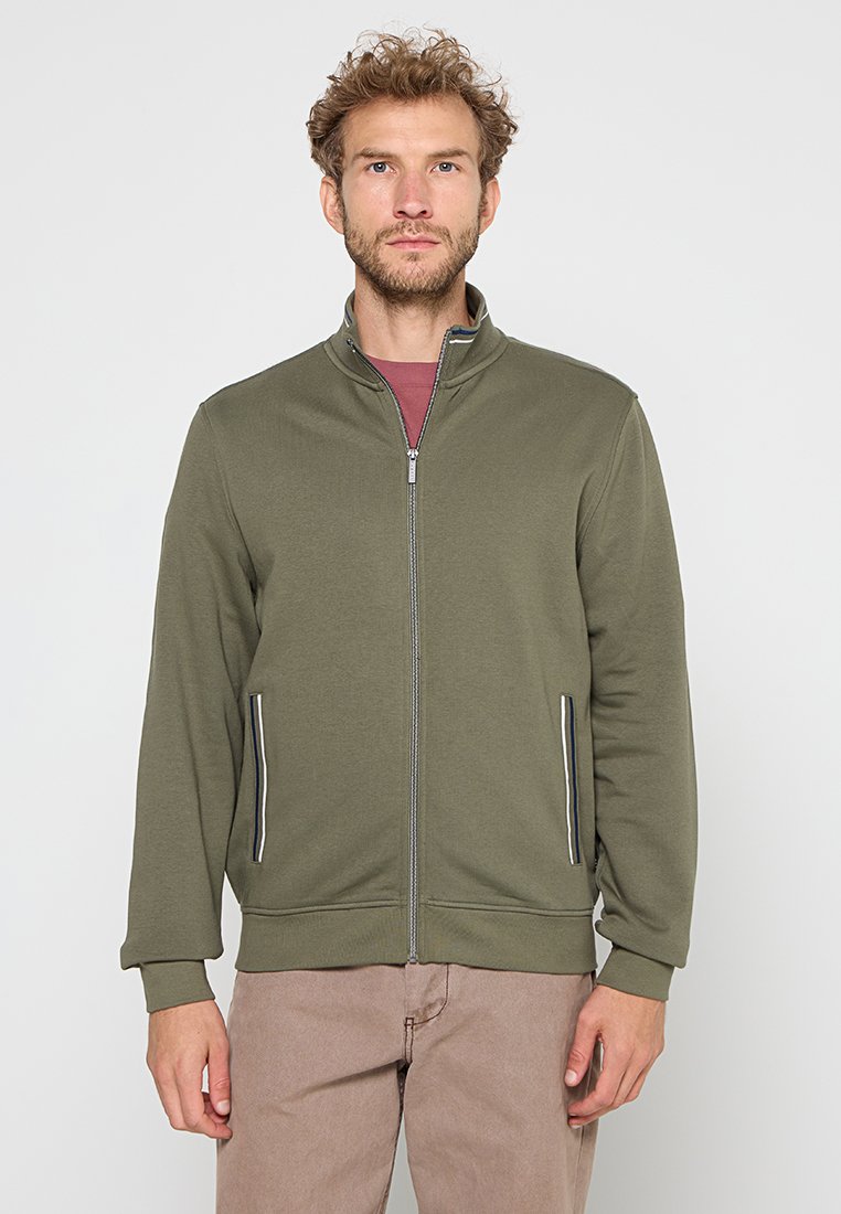 Uomo che indossa una giacca verde oliva con zip con accenti a strisce sulle tasche e pantaloni marrone chiaro, su uno sfondo semplice.