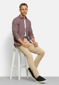 Männliches bordeauxrotes Button-Up-Hemd mit einer Tasche, über einem weißen T-Shirt getragen. Beigefarbene Hose und schwarze Sneakers vervollständigen das Outfit. Sitzt auf einem Hocker.