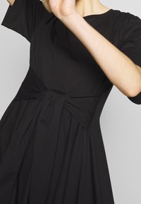 Kvinna klädd i en svart klänning med korta ärmar och detaljerad midja, som lyfter en arm framför en neutral bakgrund.