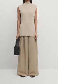 Beżowy, prążkowany top bez rękawów w zestawieniu z szerokimi, beżowymi spodniami, trzymając czarną, plecioną torebkę. Gładkie tekstury, neutralna paleta kolorów.
