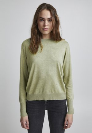 Pull vert clair en tissu doux, avec un col rond et des poignets côtelés, associé à un jean gris foncé.