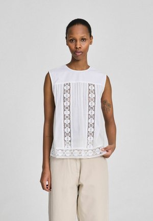 Woman wearing sleeveless white blouse with floral lace panels and beige wide-leg pants, standing against plain gray background.