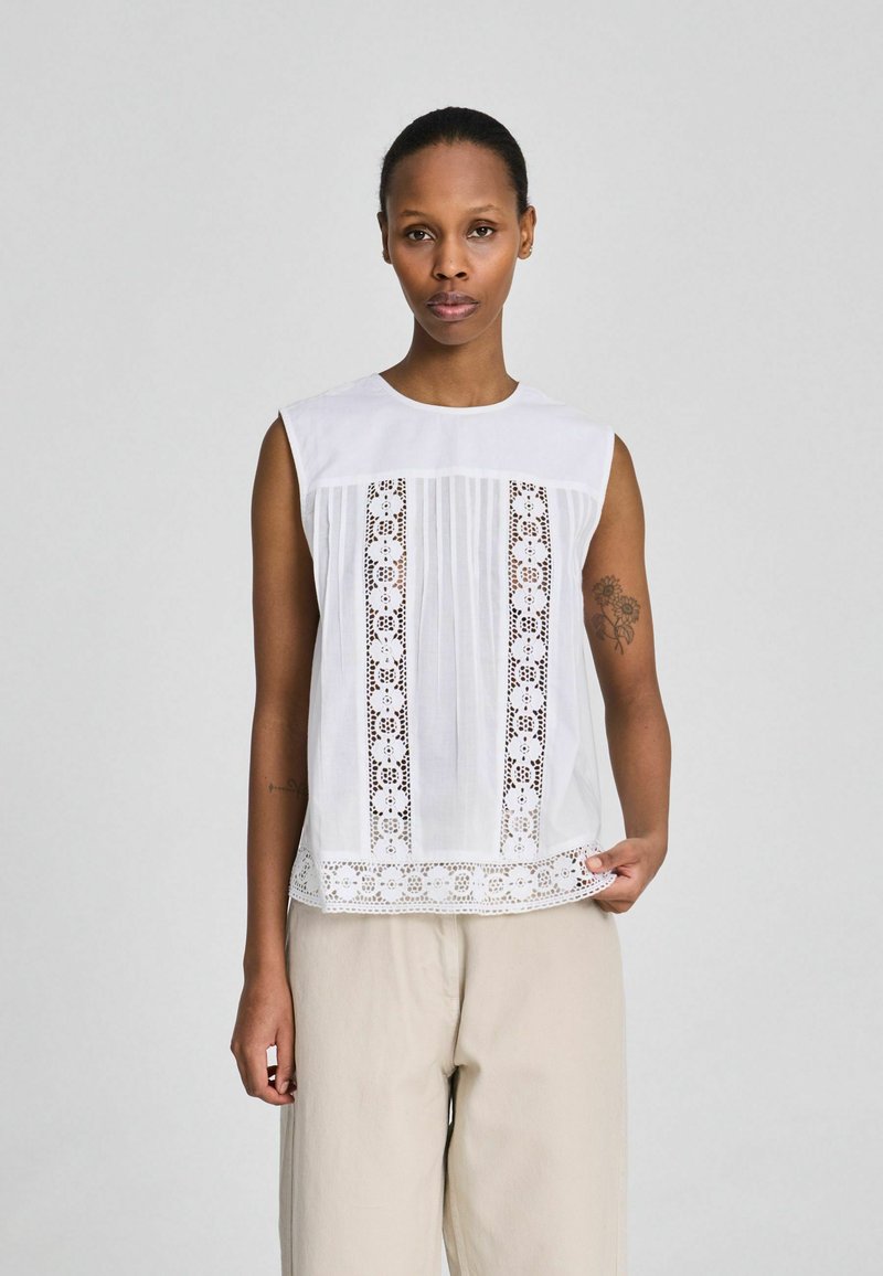 Nő ujjatlan fehér blúzban, amelyen virágos csipkepanelek vannak, és bézs színű, bőszárú nadrágot visel, szürke egyszínű háttér előtt áll.