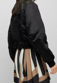 Svart satin bomberjacka med samlad nederkant och manschetter, tillsammans med en flerfärgad mönstrad kjol med djärva geometriska former.