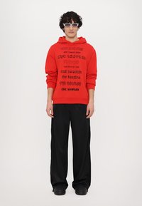 Rode hoodie met zwarte grafische tekst, gecombineerd met losse zwarte broeken en robuuste zwarte schoenen. Model draagt rechthoekige bril.
