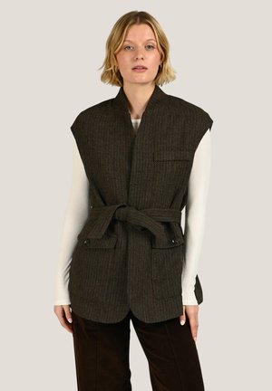 Femme portant un gilet sans manches marron foncé en chevrons avec une taille ceinturée, par-dessus un haut blanc à manches longues et un pantalon foncé, debout sur un fond beige.