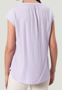 Blouse lavande clair avec un col rond, des manches courtes et un détail plissé au dos. Texture douce et lisse ; ourlet légèrement plus long à l'arrière.