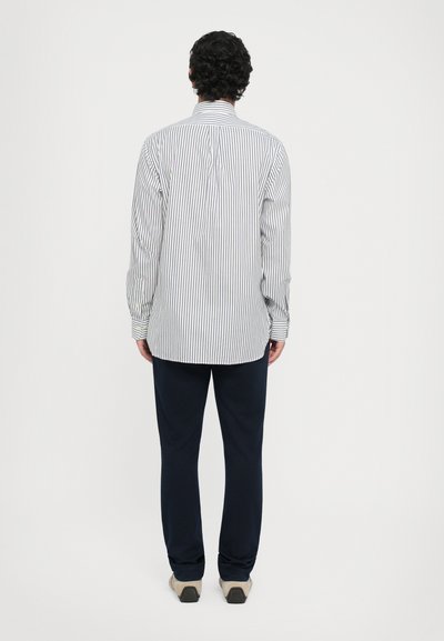 Chemise à rayures à manches longues blanche et bleu marine, avec un col boutonné et une coupe droite, associée à un pantalon bleu marine et des chaussures beiges.