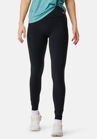 Legging noirs avec une texture lisse et extensible, dotés de poches latérales, d'une taille haute et d'une coupe ajustée. Des baskets blanches sont portées.