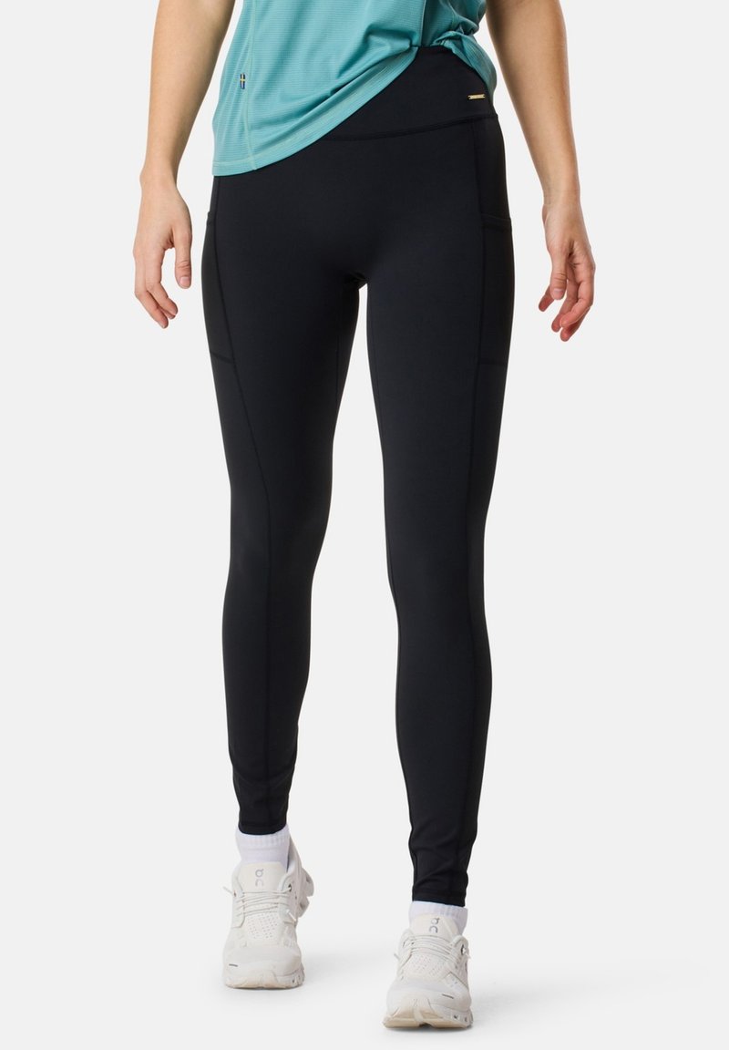 Legging noirs avec une texture lisse et extensible, dotés de poches latérales, d'une taille haute et d'une coupe ajustée. Des baskets blanches sont portées.