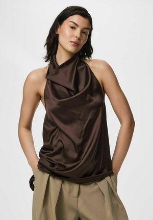 Femme portant un haut dos nu en satin marron foncé avec un pantalon plissé beige, debout les mains dans les poches devant un fond uni.