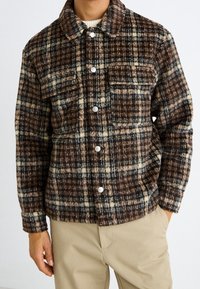 Mężczyzna ubrany w brązową, beżową i czarną koszulę w kratę z kieszeniami na piersi, noszącą białe swetry, w połączeniu z beżowymi spodniami.