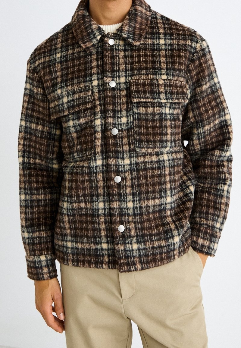 Mężczyzna ubrany w brązową, beżową i czarną koszulę w kratę z kieszeniami na piersi, noszącą białe swetry, w połączeniu z beżowymi spodniami.