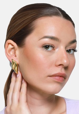 Donna con pelle liscia e capelli castani che indossa grandi orecchini dorati, lucidi e scultorei, toccandosi un lobo dell'orecchio, guarda leggermente di lato.