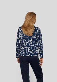 Dunkelblauer Pullover mit weißen abstrakten Mustern, langen Ärmeln und rundem Ausschnitt. Strukturierter Stoff mit lockerer Passform. Getragen mit dunklen Hosen.