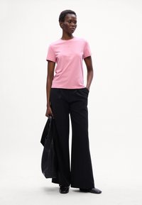 Pink bomulds T-shirt med korte ærmer parret med højtaljede sorte brede bukser. Modellen holder en sort tote-bag med en glat tekstur.