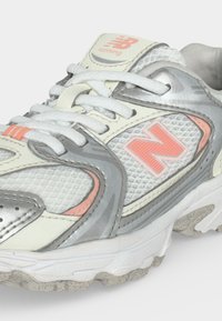 Λευκό και γκρι παπούτσι τρεξίματος New Balance με κοραλί λεπτομέρειες, υφή από δίχτυ και λευκά κορδόνια.