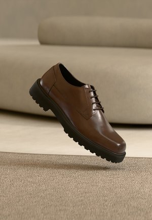 Pantof din piele maro tip oxford, cu talpă neagră groasă, plutind deasupra unui covor bej texturat în fața unei canapele deschise la culoare.