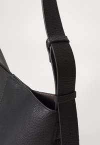 Lanière de sac à main en cuir noir texturé avec boucle ajustable et partie du corps du sac visible sur un fond uni.