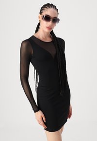 Rochie mini neagră cu corset ajustat, mâneci lungi transparente și decolteu în V. Modelul poartă ochelari de soare supradimensionați; părul este coafat în împletituri.