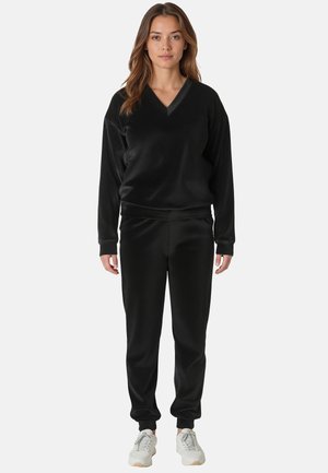 Schwarzes Sportset bestehend aus einem langärmeligen V-Ausschnitt-Oberteil und tapered Hosen, aus glattem Material gefertigt. Kombiniert mit leichten Sneakers.