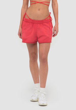 Kvinde iført koralrøde shorts med snor og matchende top med stropper samt hvide sokker og sneakers, stående mod en ensfarvet baggrund.