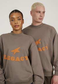 Hanorac maro cu grafica imprimată portocalie, inclusiv stele și cuvântul "LEGACY". Are un guler înalt și o croială lejeră.