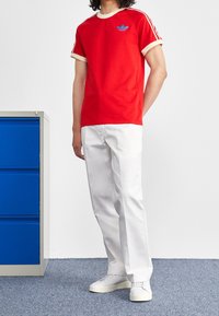 Červené bavlněné tričko s krémovým lemováním a modrým logem, v kombinaci s bílými kalhotami a světlými teniskami, stojící vedle modré skříně.