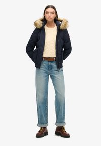 Doudoune marine avec capuche en fausse fourrure, pull en tricot crème à torsades, jeans bleu clair à jambes larges et bottes brunes à lacets.