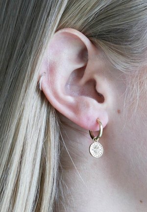 Gouden hoepel oorbellen met een ronde hanger die een zonnestraalontwerp heeft. De oorbellen hangen dicht bij het oorlelletje en tonen een gladde textuur.
