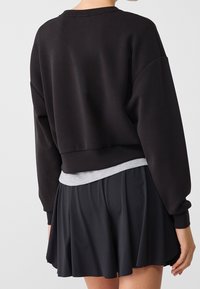 Sweat-shirt noir raccourci à manches longues, associé à une ceinture grise et une jupe noire évasée, offrant un look décontracté en superposition.