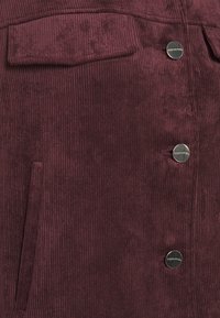 Tissu en velours côtelé bordeaux profond avec trois boutons métalliques étiquetés "comma", une poche à rabat et une ouverture de poche latérale verticale.