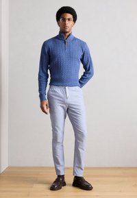 Μπλε πουλόβερ με πλέξη καλσόν και γιακά με φερμουάρ μέχρι τη μέση, συνδυασμένο με ανοιχτό μπλε παντελόνι και σκούρα καφέ μοκασίνια, στέκεται σε ξύλινο πάτωμα.