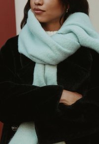 Sciarpa azzurro chiaro, misto lana, avvolta attorno al collo. Abbinata a un cappotto plush nero. Texture morbida, che evidenzia un'estetica accogliente.