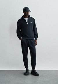 Μαύρη φούτερ με φερμουάρ και λογότυπο, συνδυασμένη με μαύρα παντελόνια joggers, παρέα με μαύρα αθλητικά παπούτσια και καπέλο, σε γκρίζο φόντο.