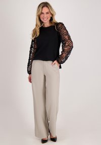 Zwarte top met doorschijnende mouwen met patroon, gecombineerd met lichtbeige broek met wijde pijpen. Zwarte puntige hakken maken de outfit compleet.