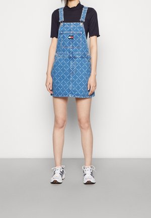 Person iført en blå mønstret denim overallkjole over en sort kortærmet top og hvide sneakers, stående mod en ensfarvet baggrund.