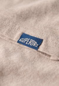 Tan stoftekstur med et blåt mærke, der læser "SUPERDRY" syet i hvidt. Nært billede, der viser blødheden og syningsdetaljerne.