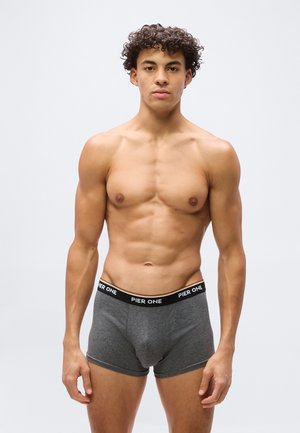 Jeune homme aux cheveux bouclés debout, portant un boxer gris avec l'inscription "PIER ONE" sur la ceinture, sur un fond clair uni.