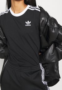 Černé tričko s bílým límcem a třemi bílými pruhy na ramenou, s malým bílým logem Adidas na prsou. Nosené pod černou koženou bundou.