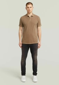 Νεαρός άντρας που στέκεται με ουδέτερη έκφραση, φοράει καφέ πόλο μπλουζάκι, πιο ανοιχτά μαύρα τζην κα�ι λευκά αθλητικά παπούτσια, σε απλό φόντο.