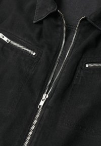 Veste en denim noire avec fermeture éclair métallique argentée et deux poches poitrine zippées horizontales, posée à plat montrant le col et les détails avant.