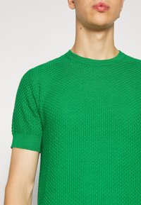 Grön stickad kortärmad tröja med ett strukturerat mönster, rund halsringning och ribbade muddar. Tyget verkar vara lätt och luftigt.