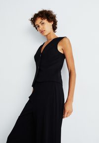 Gilet sans manches noir avec un décolleté en V profond, présentant deux boutons sur le devant. Fabriqué à partir d'un tissu lisse, avec une coupe ajustée et un design minimaliste.