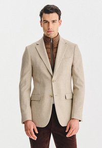 Beżowa żebrowana marynarka z dwoma przednimi kieszeniami, zapinana na jeden guzik; noszona na brązowej pikowanej kurtce z zamkiem; ciemnobrązowe spodnie.